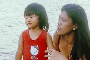 A young child wearing a red tank top with a cartoon character print stands near a body of water. An adult with long hair, wearing a similar red outfit, is next to the child, gently holding them and gazing in their direction. The water in the background is calm and expansive.