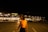 A dimly lit parking lot at night with a large, illuminated building in the background displaying the word 'METRO' on the storefront. A person wearing a bright orange top is slightly blurred in the foreground, walking towards the right side of the image.