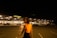 A dimly lit parking lot at night with a large, illuminated building in the background displaying the word 'METRO' on the storefront. A person wearing a bright orange top is slightly blurred in the foreground, walking towards the right side of the image.