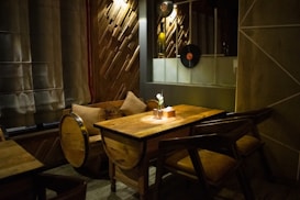 A warm and cozy dining area with a wooden table and matching chairs, illuminated by soft, dim lighting. The room has a rustic feel with wooden paneling on the walls and a window with sheer curtains. A small vase with a single flower sits atop the table, adding a simple decorative touch. A vintage vinyl record is mounted on the wall, enhancing the retro ambiance.