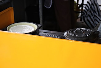 A yellow divider partially conceals a cooking setup with a black pot next to a pan containing a white batter or dough. The background is dimly lit, with a striped fabric visible.