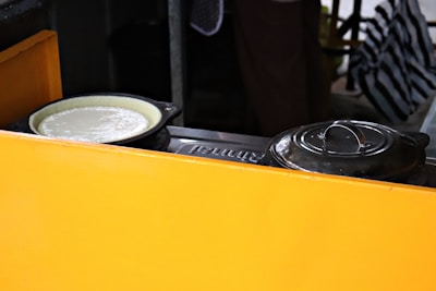 A yellow divider partially conceals a cooking setup with a black pot next to a pan containing a white batter or dough. The background is dimly lit, with a striped fabric visible.