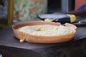 A round, crispy-edged cake or pastry with a soft, creamy filling sits on a wooden surface. The background is slightly blurred, showing part of a kitchen utensil, possibly a grater, with a yellow handle.