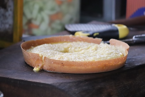 A round, crispy-edged cake or pastry with a soft, creamy filling sits on a wooden surface. The background is slightly blurred, showing part of a kitchen utensil, possibly a grater, with a yellow handle.