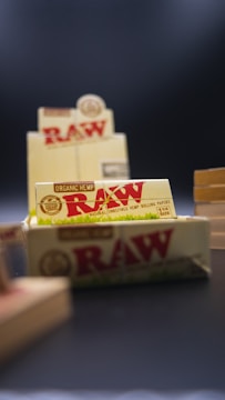 Several packages of RAW organic hemp rolling papers are prominently displayed. The focus is on a box in the foreground containing smaller packages, with the distinctive red and brown RAW branding clearly visible. Other similar packages are slightly blurred in the background, and the image overall has a shallow depth of field.