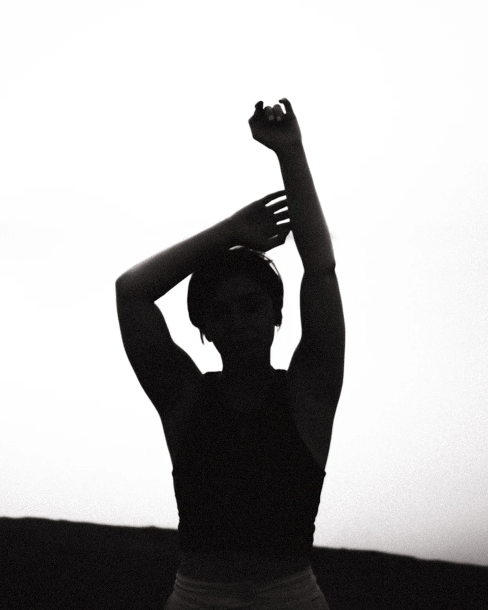 Person stands with arms raised high above head, hands on shoulders in stark contrast