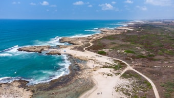 A coastal landscape with an expansive view of the ocean meeting the shoreline. The water is a vibrant turquoise, and there are rocky outcrops interspersed along the edge. The land adjacent to the coast is a mix of sandy beaches and green patches, with a dirt path winding through it. A few small structures or umbrellas are visible near the shoreline, and a couple of people can also be seen walking along the path.