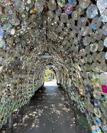 An archway made entirely of reflective CDs creates a tunnel effect, with sunlight filtering through and casting patterns of light and shadow on the ground. Inside, the atmosphere feels surreal and futuristic, with vibrant reflections and iridescent colors. There is another figure in the distance, partially visible outside the tunnel, enjoying the sunny day in what seems to be a garden or park setting.