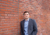 A person in business attire stands against a red brick wall. The individual is wearing a blue button-up shirt under a dark gray suit jacket. The background features a textured and slightly weathered brick surface.