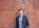 A person in business attire stands against a red brick wall. The individual is wearing a blue button-up shirt under a dark gray suit jacket. The background features a textured and slightly weathered brick surface.