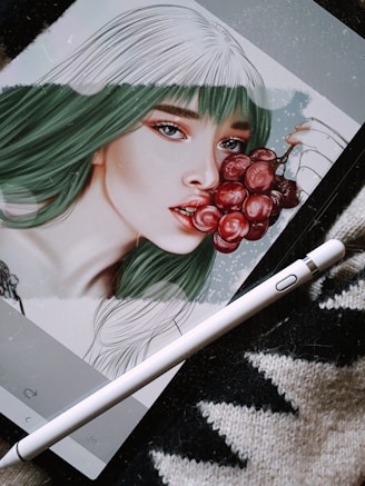 A digital artwork displayed on a tablet depicts a stylized portrait of a woman with green and white hair, holding a bunch of red grapes near her lips. The image is accompanied by a digital pen resting on a textured surface, suggesting the setting is an artistic workspace.