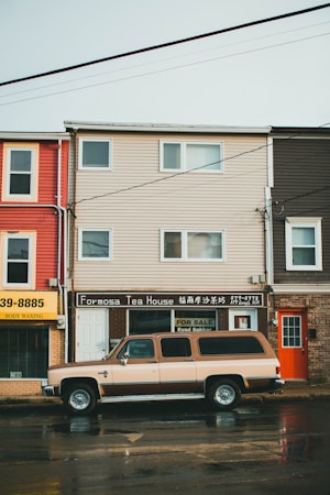 A street view featuring a tea house named Formosa Tea House with signage in both English and Chinese. A large, vintage-style SUV is parked in front of the building. The surroundings include neighboring buildings with bright colors, such as red and yellow. The street appears wet, suggesting recent rain. Power lines cross the sky above.