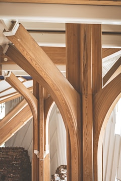 Elegant wooden beams with a curved design create a visually striking architectural feature. The light woodgrain contrasts with the white ceiling and walls, lending a natural and sophisticated ambiance to the space. In the background, there is a stack of bricks partially visible.