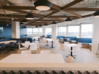 A modern, spacious cafeteria or break room with large windows allowing natural light to fill the space. The room features multiple tables with white and black chairs, blue booth seating along the windows, and a wooden ceiling grid with black pendant lights. Neutral colors and minimalist design elements create a clean and inviting atmosphere.