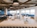 A modern, spacious cafeteria or break room with large windows allowing natural light to fill the space. The room features multiple tables with white and black chairs, blue booth seating along the windows, and a wooden ceiling grid with black pendant lights. Neutral colors and minimalist design elements create a clean and inviting atmosphere.