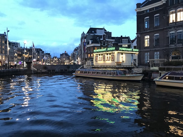 A canal scene at dusk with a brightly lit boat and surrounding buildings. The boat displays the sign 'Rondvaart Kooy' and is docked along the canal. The water reflects the lights from the boat and nearby buildings, creating a shimmering effect. The buildings are typical of European architecture, with multiple stories and ornate facades.