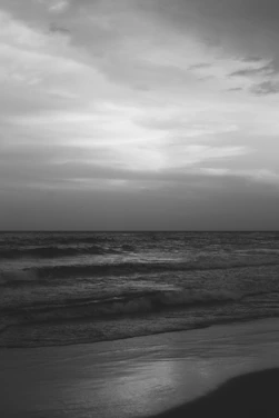 A serene black and white beach scene with waves gently rolling under a cloudy sky.