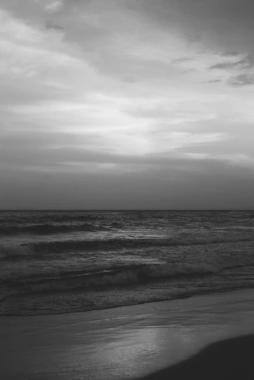 A serene black and white beach scene with waves gently rolling under a cloudy sky.