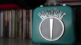 A teal-colored retro-style speaker with the brand name 'HONEYTONE' in metallic lettering on top. It has a classic grille in the center and is placed on a shelf with a collection of books or CDs blurred in the background.