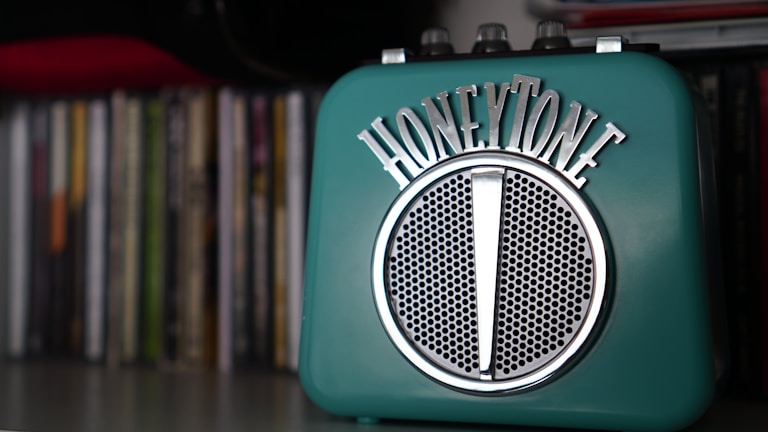 A teal-colored retro-style speaker with the brand name 'HONEYTONE' in metallic lettering on top. It has a classic grille in the center and is placed on a shelf with a collection of books or CDs blurred in the background.