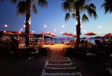 A serene beachside setting at dusk with strings of lights illuminating the area. Palm trees are prominently placed on both sides, casting soft shadows. People are seated at various tables, enjoying the setting under straw umbrellas, while the sandy path leads towards the calm sea.