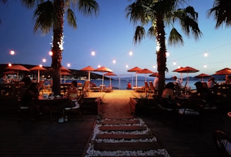 A serene beachside setting at dusk with strings of lights illuminating the area. Palm trees are prominently placed on both sides, casting soft shadows. People are seated at various tables, enjoying the setting under straw umbrellas, while the sandy path leads towards the calm sea.