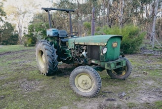 A vintage green tractor with visible wear stands in a rural outdoor setting with dirt and sparse vegetation in the background. The tractor has a John Deere logo and is parked on uneven ground, surrounded by trees and bushes.