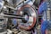 A complex industrial setup with a large circular machine at the center, surrounded by numerous cables of various colors, primarily blue and black. The machinery is metallic with a shiny texture and intricate panels. The environment appears highly technical, resembling a laboratory or a facility handling high-tech equipment.