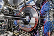 A complex industrial setup with a large circular machine at the center, surrounded by numerous cables of various colors, primarily blue and black. The machinery is metallic with a shiny texture and intricate panels. The environment appears highly technical, resembling a laboratory or a facility handling high-tech equipment.