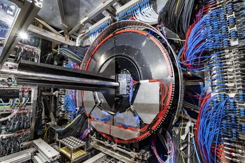 A complex industrial setup with a large circular machine at the center, surrounded by numerous cables of various colors, primarily blue and black. The machinery is metallic with a shiny texture and intricate panels. The environment appears highly technical, resembling a laboratory or a facility handling high-tech equipment.