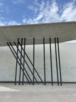 A modern architectural design featuring a concrete roof supported by sleek, black metal beams angled artistically against a textured concrete wall. The sky above is partly cloudy with patches of blue.