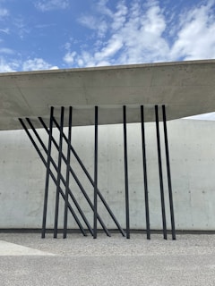 A modern architectural design featuring a concrete roof supported by sleek, black metal beams angled artistically against a textured concrete wall. The sky above is partly cloudy with patches of blue.