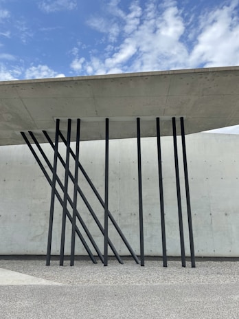 A modern architectural design featuring a concrete roof supported by sleek, black metal beams angled artistically against a textured concrete wall. The sky above is partly cloudy with patches of blue.