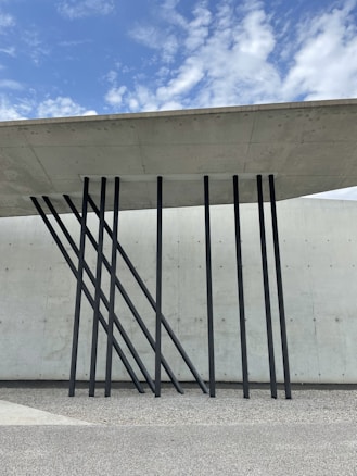A modern architectural design featuring a concrete roof supported by sleek, black metal beams angled artistically against a textured concrete wall. The sky above is partly cloudy with patches of blue.