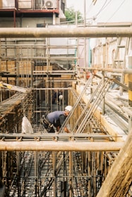 A worker wearing a hard hat is bending over to work on a construction site filled with metal beams and scaffolding. The setting appears industrial with complex network of metal rods and wooden structures. In the background, there are residential buildings and outdoor cables.