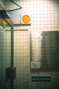 A tiled wall featuring a directory sign listing various business names and room numbers in a building. There is a security camera sign with an orange logo attached above. The setting appears to be an indoor corridor or lobby area with a glass door on the left side.