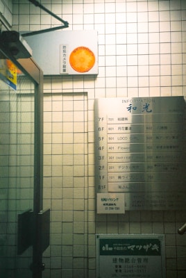A tiled wall featuring a directory sign listing various business names and room numbers in a building. There is a security camera sign with an orange logo attached above. The setting appears to be an indoor corridor or lobby area with a glass door on the left side.