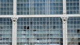 A section of a large window comprising multiple glass panes arranged in a grid pattern. Some of the panes are broken or missing. The window is framed by white pillars featuring ornate decorative designs at the top.