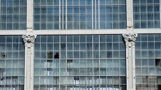 A section of a large window comprising multiple glass panes arranged in a grid pattern. Some of the panes are broken or missing. The window is framed by white pillars featuring ornate decorative designs at the top.