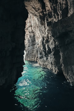 A natural rock formation forms a narrow passageway, revealing clear turquoise waters below. The sunlight filters through the opening, illuminating parts of the rocky walls and the shimmering water surface.