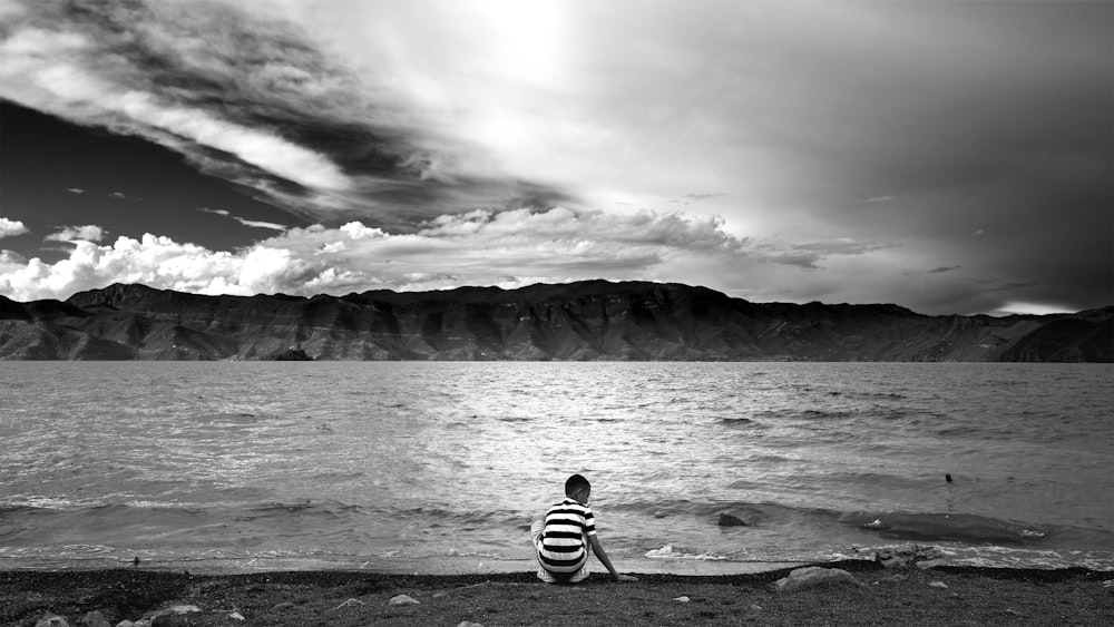grayscale photo of man in stripe shirt sitting on rock near body of water