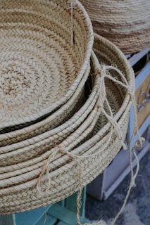 A stack of woven baskets made of natural fibers is arranged on a surface. Each basket displays intricate and textured patterns with visible strands of material. The baskets have a light tan color and there are loose fibers hanging from the edges.