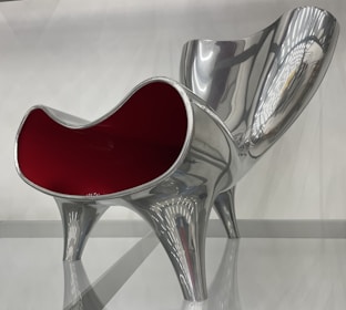 A modern and artistic chair with a glossy silver finish and a striking red interior, featuring a curvy and sculptural design. The chair has a futuristic appearance with clean lines and reflective surfaces, supported by four sturdy legs.