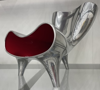 A modern and artistic chair with a glossy silver finish and a striking red interior, featuring a curvy and sculptural design. The chair has a futuristic appearance with clean lines and reflective surfaces, supported by four sturdy legs.