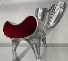 A modern and artistic chair with a glossy silver finish and a striking red interior, featuring a curvy and sculptural design. The chair has a futuristic appearance with clean lines and reflective surfaces, supported by four sturdy legs.
