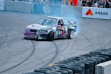 A colorful race car is drifting around a track, leaving behind a trail of smoke. The car is adorned with various sponsorship stickers and graphics. It is performing in front of a crowd, which is separated from the track by barriers. The ground shows skid marks from previous drifts.