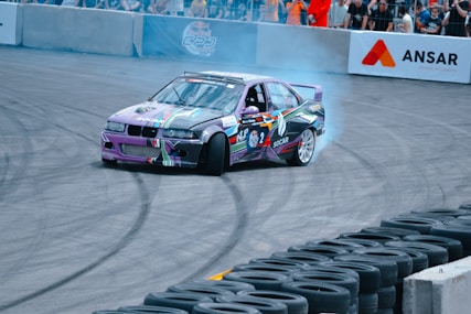 A colorful race car is drifting around a track, leaving behind a trail of smoke. The car is adorned with various sponsorship stickers and graphics. It is performing in front of a crowd, which is separated from the track by barriers. The ground shows skid marks from previous drifts.