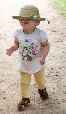 A toddler wearing a straw hat that covers most of the face, dressed in a white shirt with a cartoon image and yellow leggings. The child is walking on a sandy path, with brown sandals on.