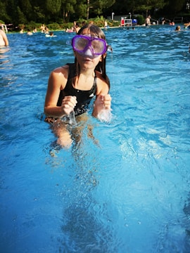 A young girl wearing a black swimsuit and purple goggles is standing in a swimming pool. She is playfully splashing water with her hands. The pool is busy, with several other people in the background enjoying the water. The surroundings are lush with trees and some poolside furniture.