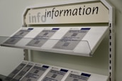A display rack filled with informational brochures is seen. The brochures have a blue and white color scheme and feature text with QR codes. Above the rack, a sign reads 'info information' in uppercase and lowercase letters.